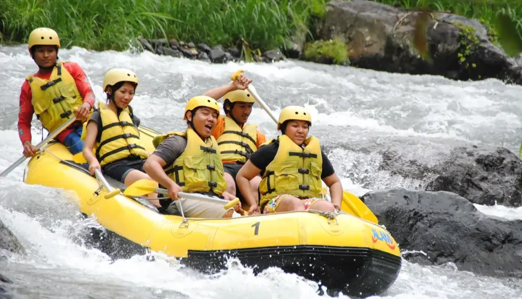 Un grupo de personas practicando rafting en un río de aguas rápidas. Están en una balsa inflable amarilla, equipados con cascos y chalecos salvavidas. La balsa está navegando a través de rápidos, y las expresiones en sus rostros muestran emoción y concentración mientras manejan los remos. La vegetación verde y las rocas son visibles en el fondo, añadiendo un toque natural al escenario.