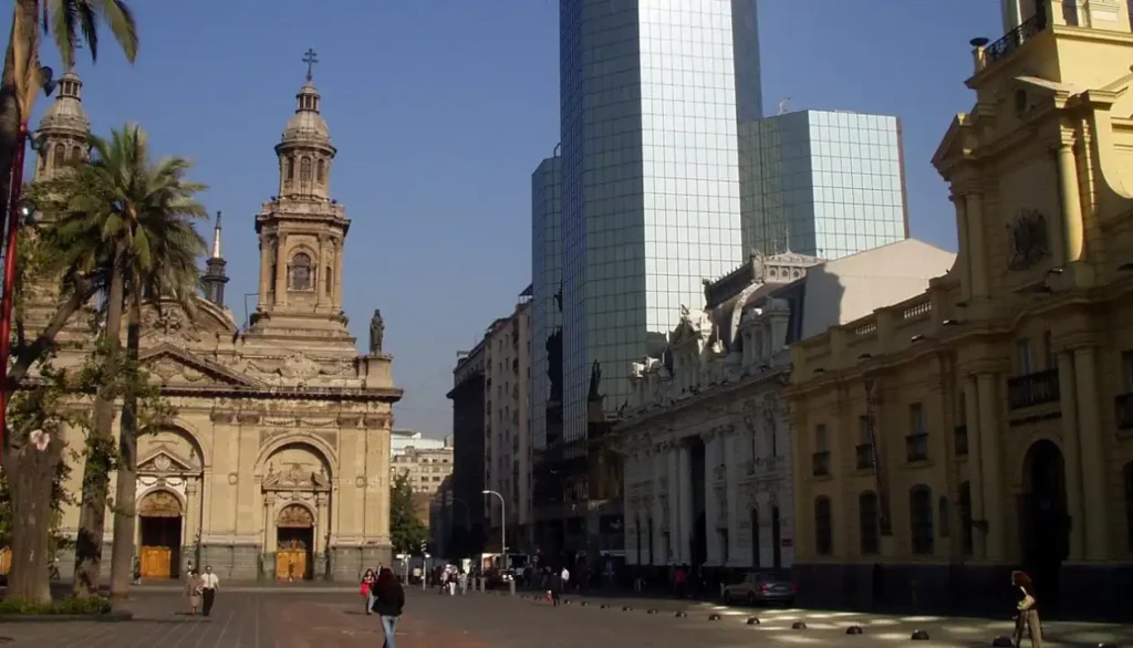 Uma vista da Plaza de Armas em Santiago. À esquerda, está a Catedral Metropolitana, uma estrutura histórica impressionante, e à direita, modernos edifícios de escritórios, representando o contraste entre o antigo e o novo na cidade.