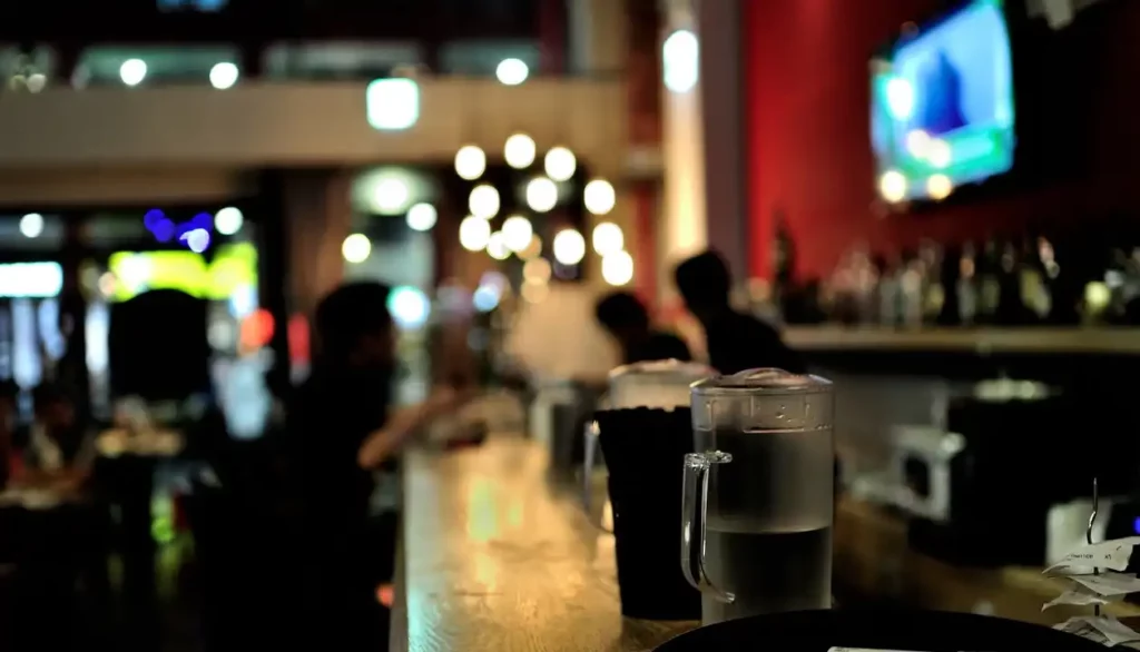 Um bar animado com iluminação suave. Em primeiro plano, há uma jarra de água sobre o balcão e, ao fundo, pessoas conversando e aproveitando o momento. As luzes pendentes e os reflexos criam um ambiente acolhedor e agradável.