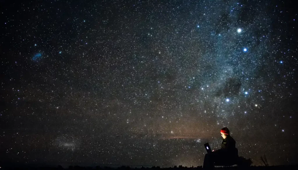 Céu noturno cheio de estrelas no Deserto do Atacama, com uma pessoa observando através de um telescópio, destacando a clareza e a beleza do céu noturno na região.