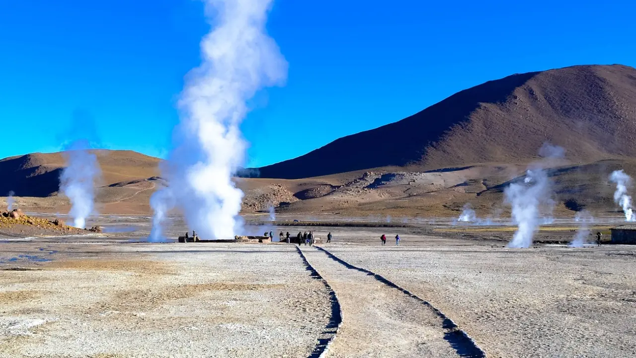 Gêiseres de vapor branco emergindo do solo na paisagem árida dos Gêiseres del Tatio no Deserto do Atacama, com montanhas ao fundo sob um céu azul claro. Turistas caminham ao longo de trilhas de terra para observar de perto o fenômeno natural.