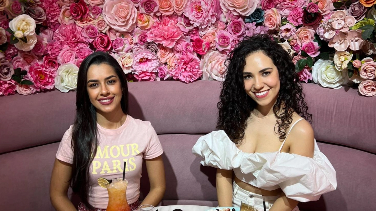 Duas amigas em uma cafeteria decorada com flores, sentadas em uma mesa de mármore com bebidas e sobremesas à sua frente, sob uma placa de neon que diz 'first coffee.