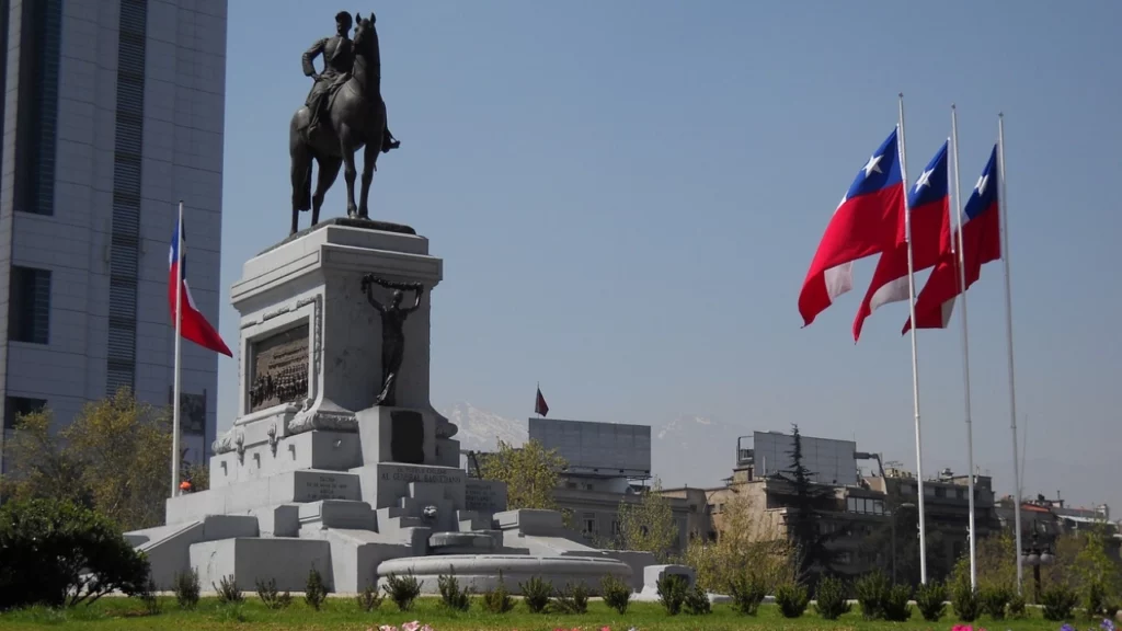 Estátua equestre em um monumento em Santiago, Chile, cercada por bandeiras chilenas e um jardim florido com edifícios modernos ao fundo