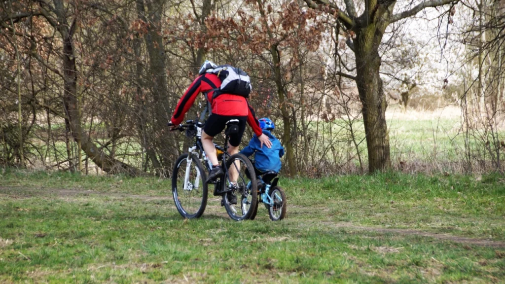 Adulto pedalando ao lado de uma criança em uma bicicleta infantil, atravessando uma trilha arborizada.