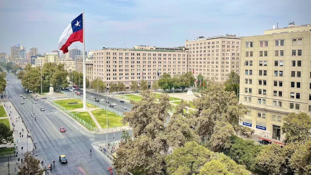 Vista aérea de uma praça em Santiago, Chile, com uma grande bandeira chilena ao centro e edifícios ao redor.