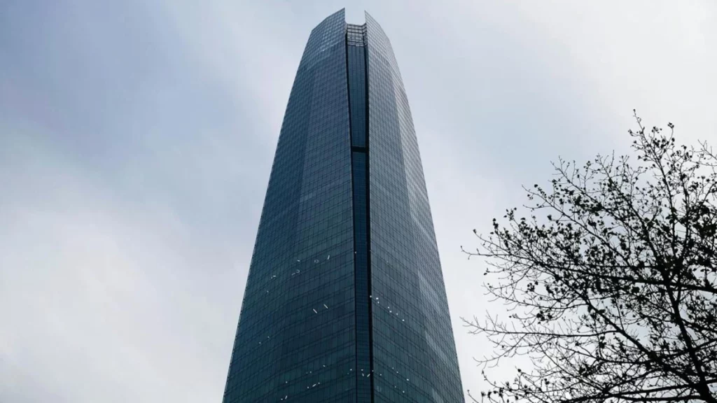 Torre Costanera, o edifício mais alto da América do Sul, em Santiago, Chile, contra um céu nublado, com árvores parcialmente visíveis em primeiro plano.