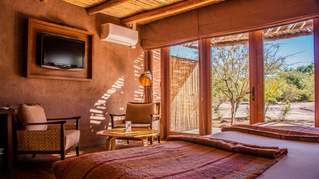 Quarto de hotel em San Pedro de Atacama com decoração rústica e elegante, mostrando uma cama de casal, janelas amplas com vista para o deserto e móveis de madeira em estilo tradicional.