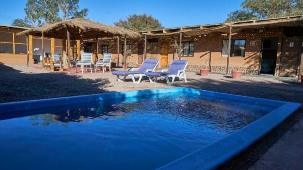 Piscina compacta em um hotel boutique de San Pedro de Atacama, com espreguiçadeiras e cabanas rústicas ao fundo, em um ambiente descontraído e ensolarado no deserto.